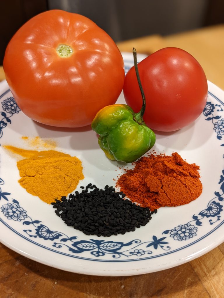 Tomatoes, scorpion pepper, chili powder, onion seeds and turmeric on a plate.